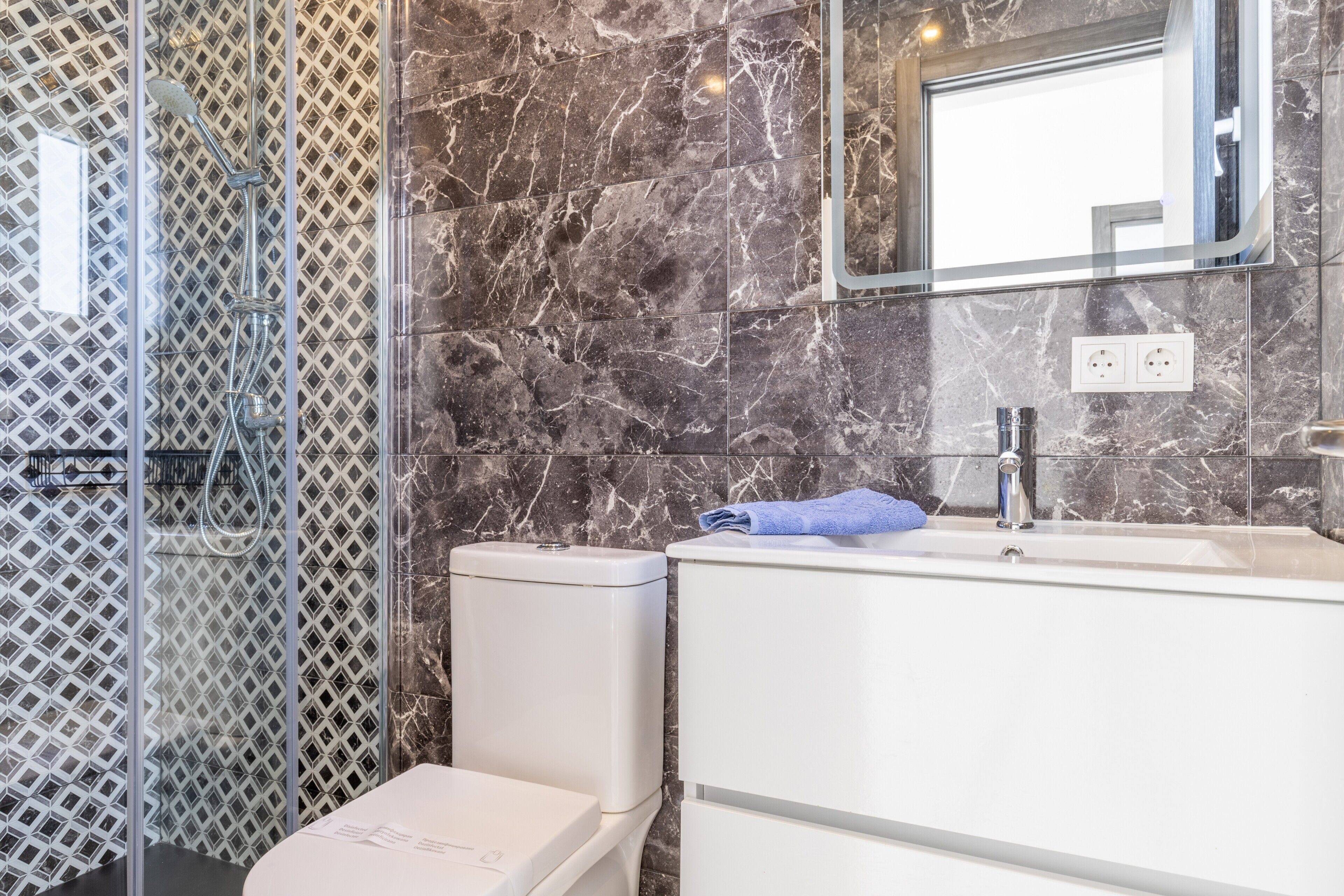 Baño con ducha, inodoro y lavamanos, que presenta paredes de mármol y una mampara de vidrio.