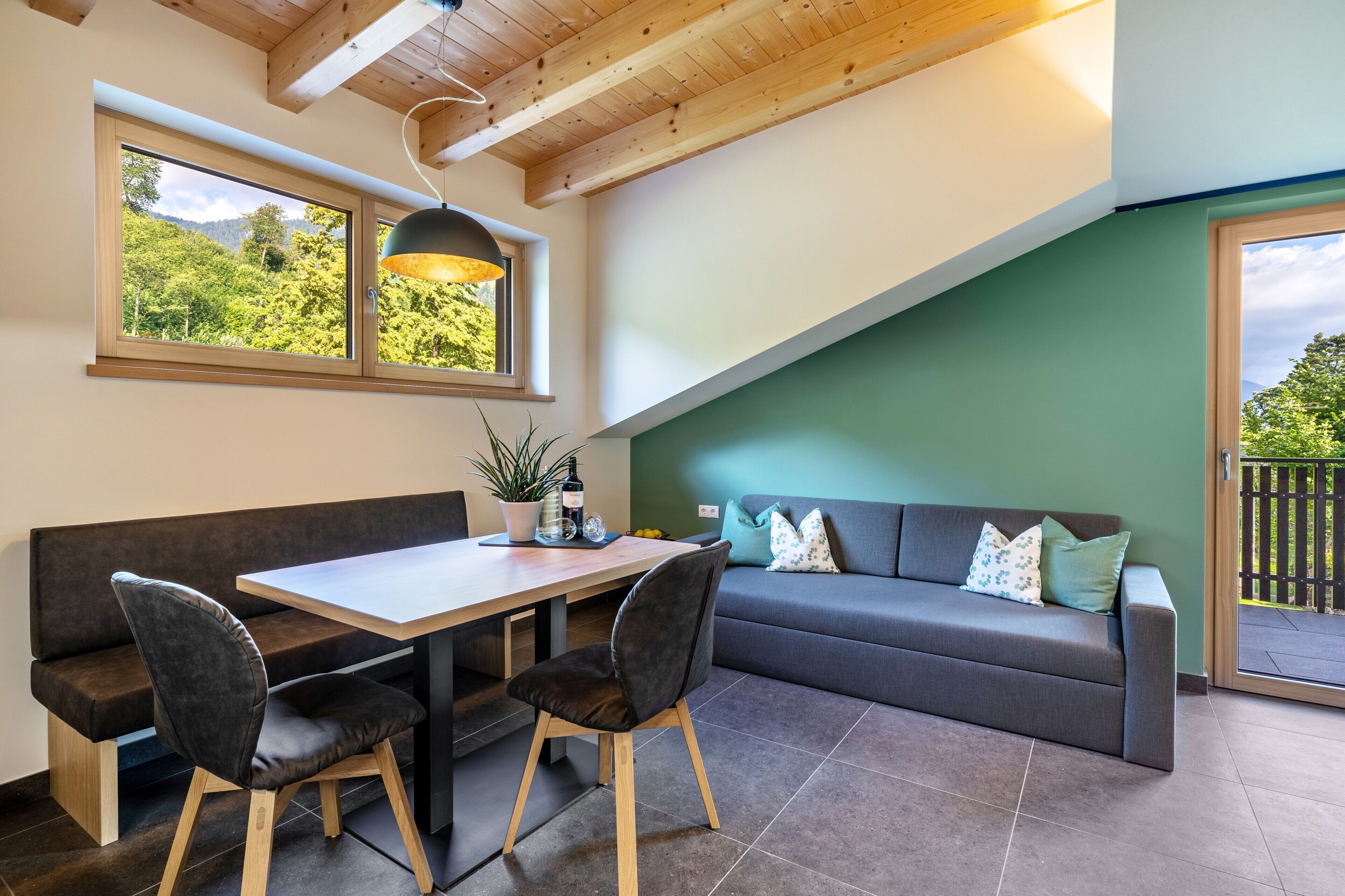 A modern interior with a wooden ceiling, a dining table with chairs, a sofa, and a view of greenery through large windows.