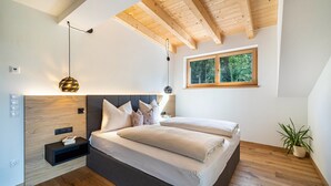 Una camera da letto moderna con soffitto in legno, un letto con molti cuscini, un comodino con una lampada e una finestra vista alberi.