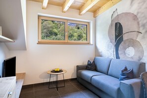 A modern living room with a grey sofa, a small round table with a bowl of fruit, a large abstract painting, and a window with a view of trees. - Apartment "Wiesgut Golden" with Mountain View, Private Terrace and Wi-Fi (Tesimo)