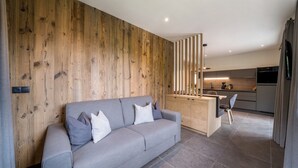 A modern living room with a grey sofa, wooden panel wall, and a kitchen in the background.