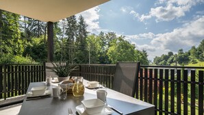 A table set for two with white dishes, a pitcher, and a potted plant on a balcony with a view of trees and a fence.