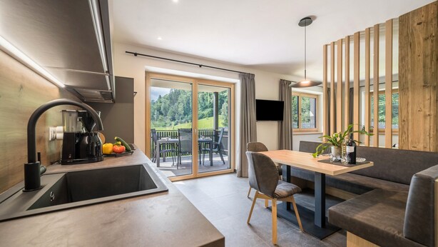 A modern kitchen with a built-in oven, a coffee machine, and a sink. There is a dining area with a table and chairs, and a sofa. The room has large windows with a view of greenery outside.