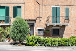 A brick building with green shutters, a small balcony, and a tree in the foreground.