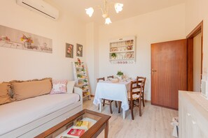 Una sala de estar con sofá, mesa de centro, mesa de comedor y sillas. Hay una estantería, un estante montado en la pared con objetos decorativos y una puerta de madera.