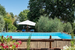 A swimming pool with a wooden deck, surrounded by lush greenery and a large umbrella.