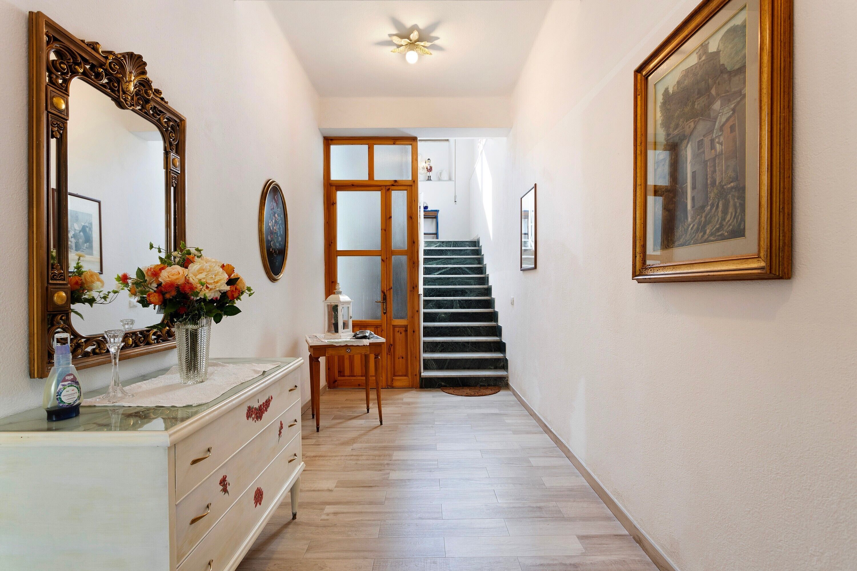 A hallway with a wooden staircase, a mirror, a vase with flowers, and a framed painting on the wall.