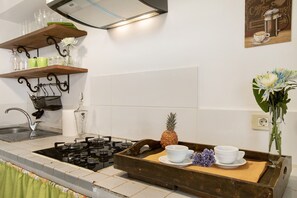 Une cuisine équipée d’une cuisinière à gaz, d’un plateau en bois avec des tasses et un ananas, et d’une étagère murale où sont rangés des verres.