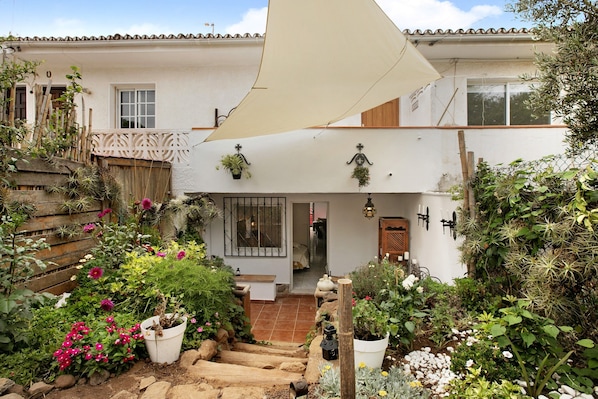 Un bâtiment blanc avec un toit recouvert de tuiles, une clôture en bois et un jardin où poussent différentes plantes et fleurs.