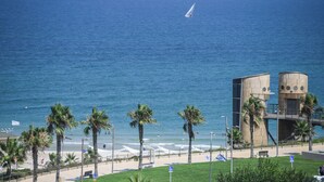 A beach view with a sailboat, palm trees, and a modern building with a unique design. - 3BR SeaView Apt by the Beach by SeaNRent (Herzliya)