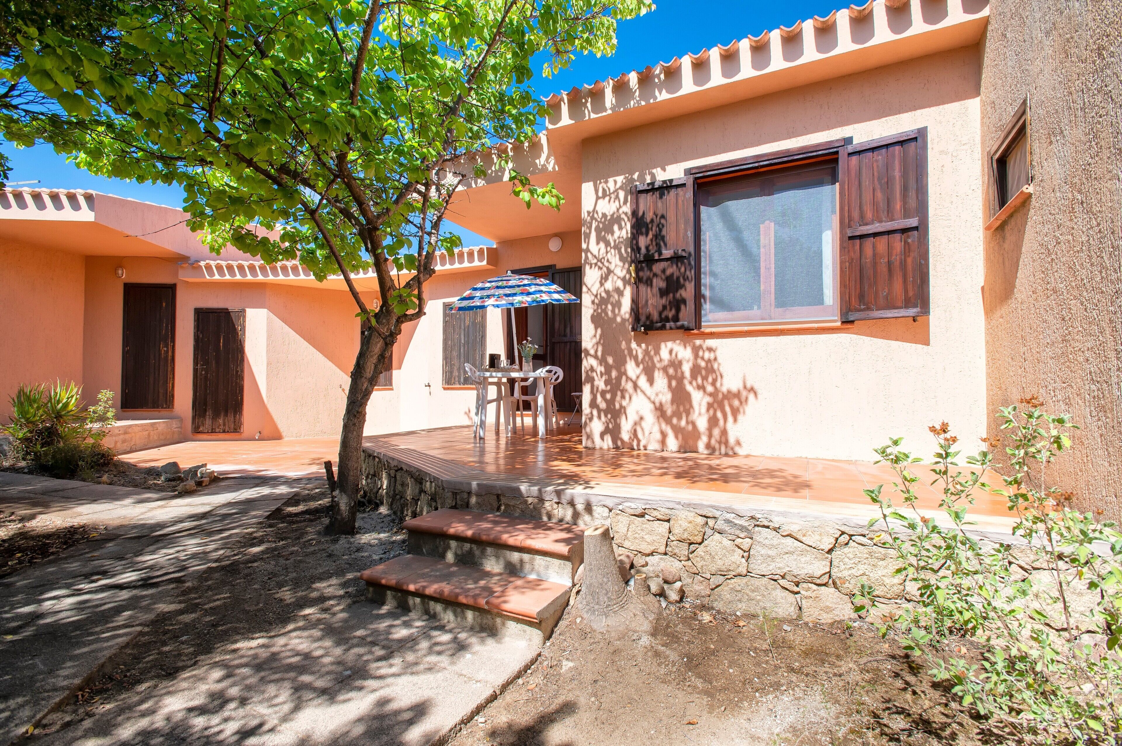 A building with a stone staircase leading to a porch, a table with chairs, and a tree providing shade.