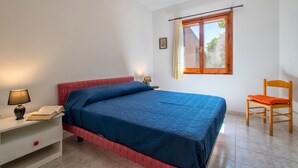 Un dormitorio con cama, mesita de noche, lámpara, silla, ventana y un cuadro en la pared.