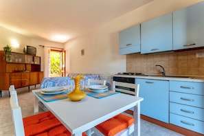 Una cucina compatta con mobili blu, un tavolo bianco con cuscini arancioni e un'area pranzo con un divano a fantasia.