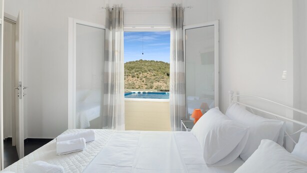 Habitación de hotel con cama, ropa blanca, vistas a la playa y montañas, y un balcón con mesa y silla.