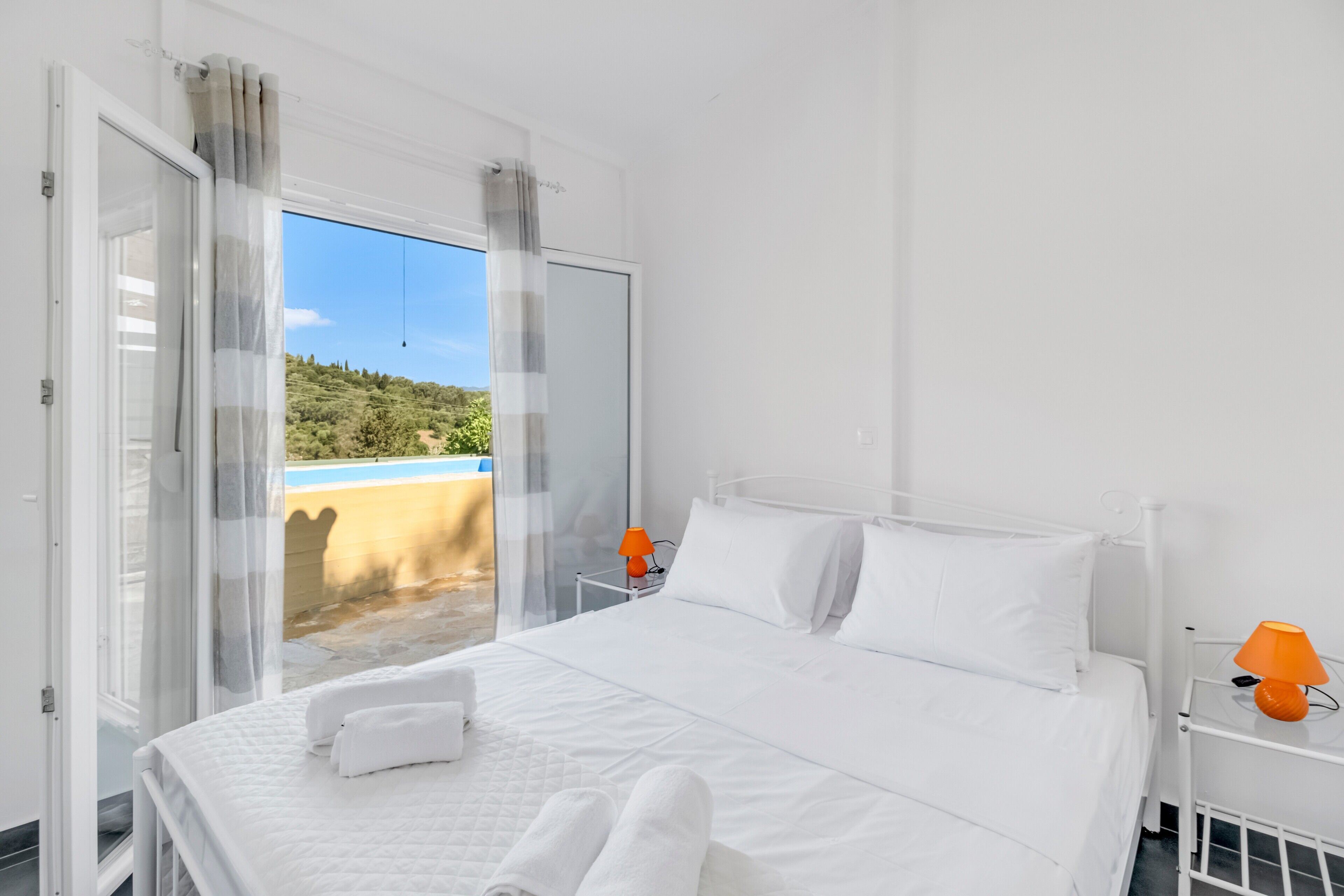 Habitación de hotel con cama, cortinas blancas, una lámpara naranja y vistas a zonas verdes.