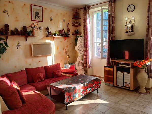 A living room with a red sofa, a coffee table with a floral tablecloth, a TV on a wooden stand, and various wall decorations.