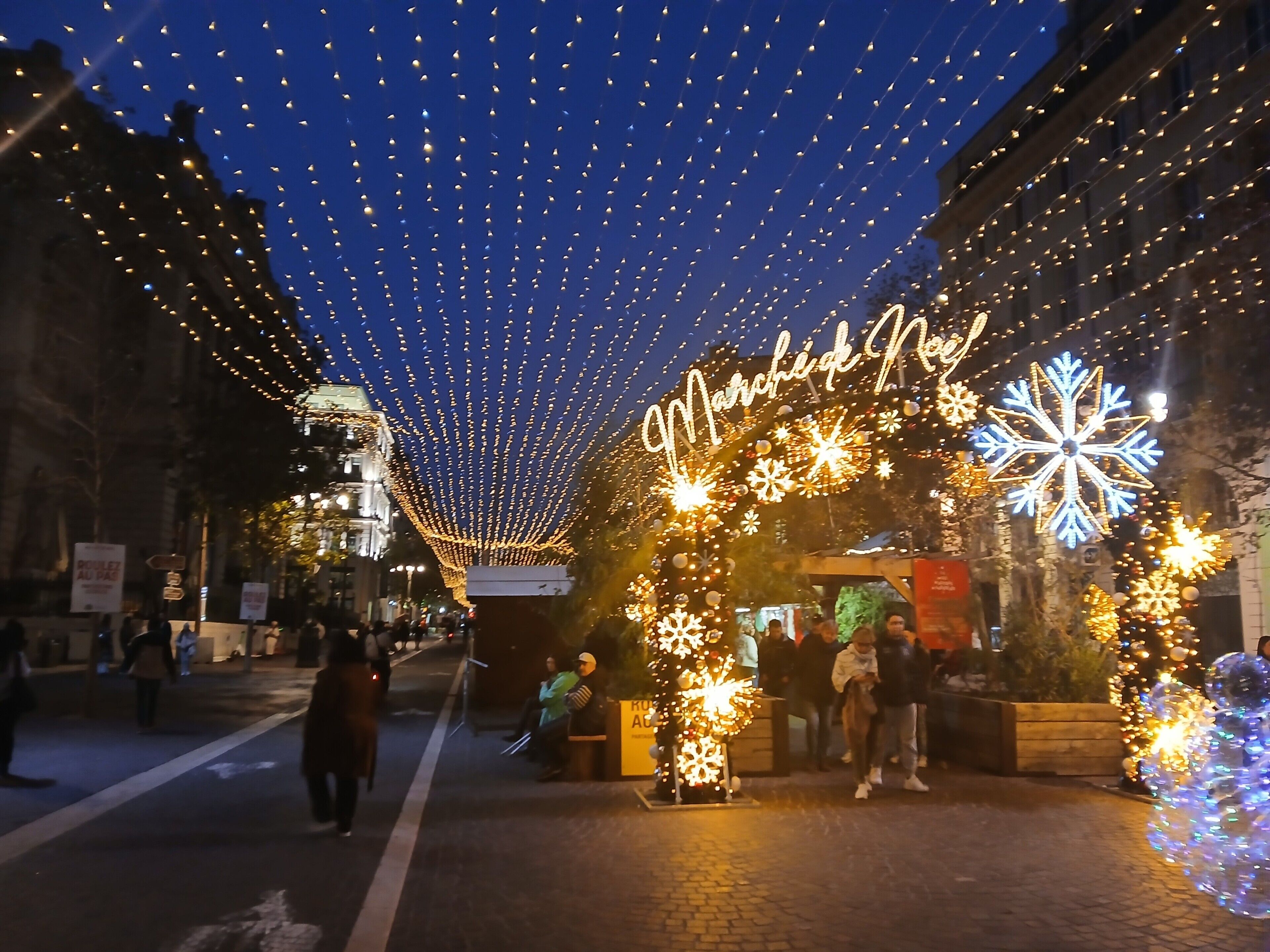 Une rue décorée de guirlandes lumineuses et de décorations en forme de flocons de neige, un banc et une pancarte indiquant « Marché de Noël ».