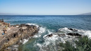 A rocky coastline with clear blue water, a few people, and a distant mountain range.