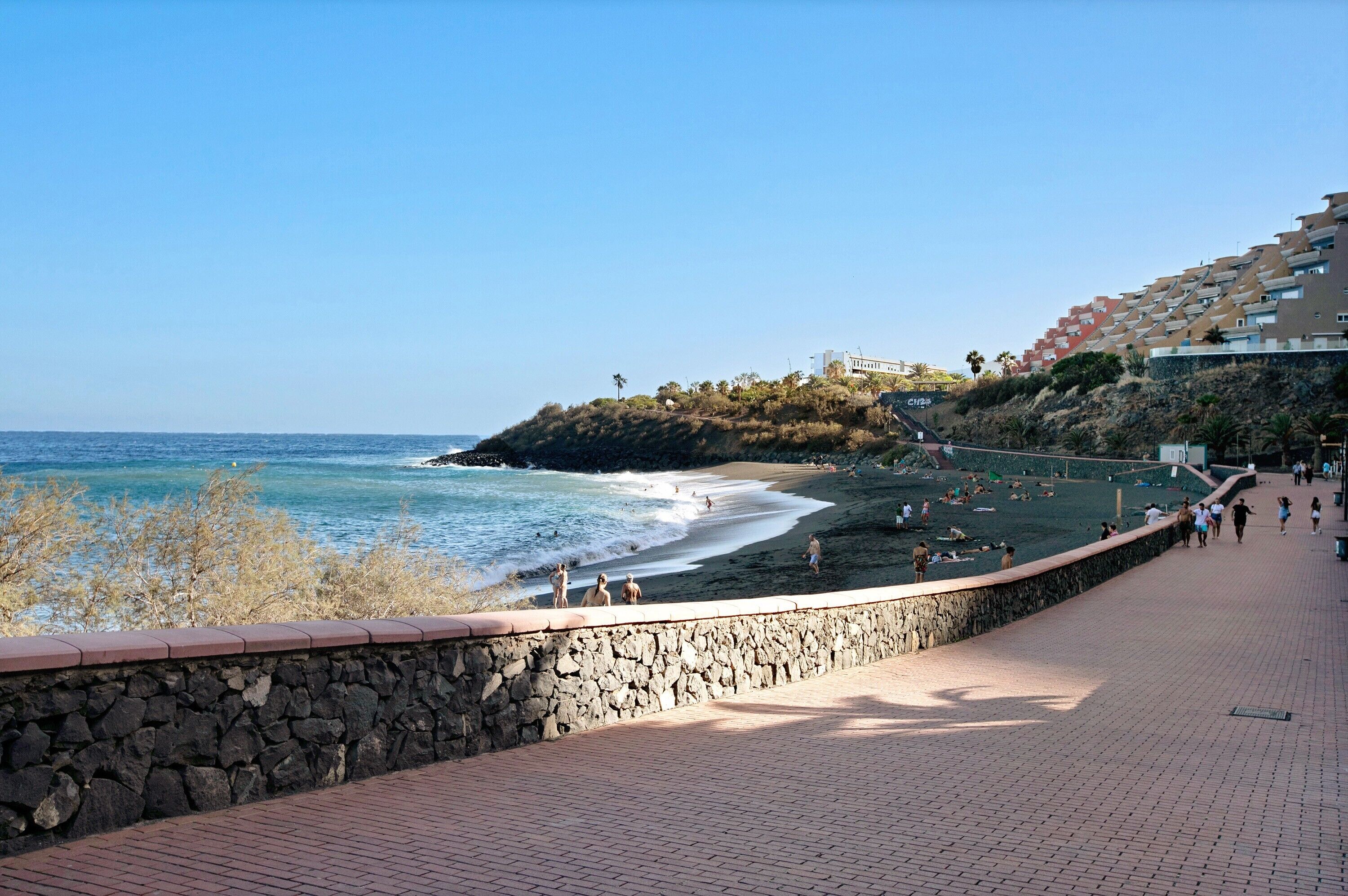 Un paseo marítimo con muro de piedra, playa y una colina con edificios.