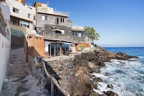 A coastal property with a stone retaining wall, a wooden deck, and a building with large windows overlooking the sea.