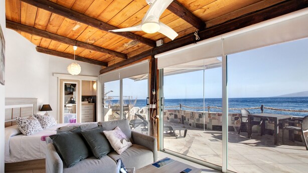 A room with a bed, a sofa, a table, and a view of the sea through large glass doors.