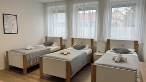 A hotel room with three single beds, each with a wooden frame and a grey bedspread. There are white curtains on the windows, and a framed picture on the wall. - Big Family Suite with 7 beds in the center of Göppingen (Göppingen)