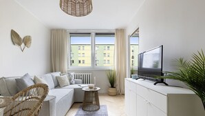 Un salon moderne avec un canapé, un téléviseur, un fauteuil en osier, une petite table d’appoint et une plante en pot.