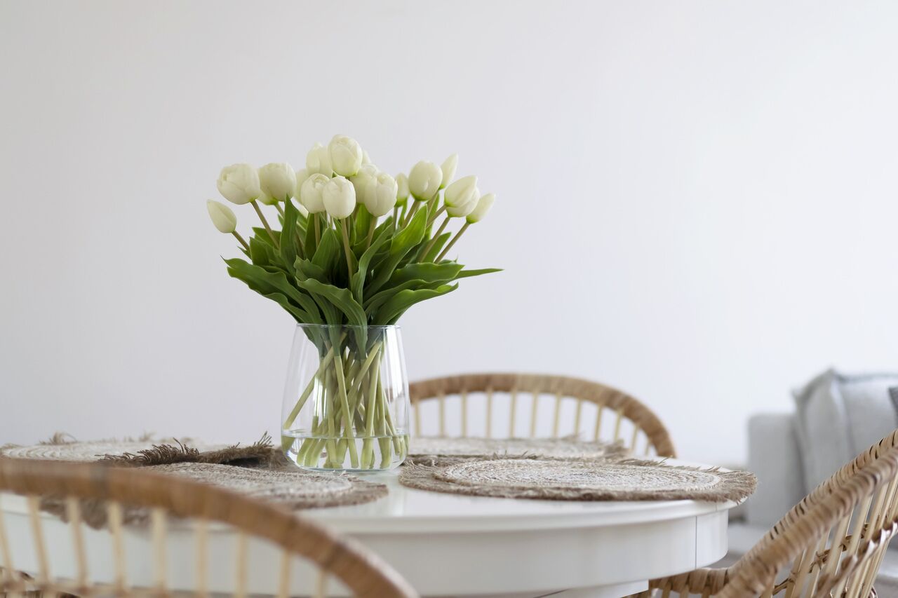 A dining table with a clear glass vase containing white tulips, surrounded by wicker chairs.