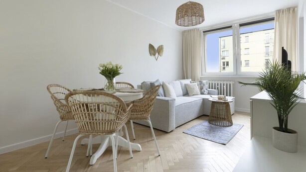 Un salon moderne avec un canapé gris clair, une table à manger entourée de chaises en osier, un luminaire suspendu et une grande fenêtre avec des rideaux.