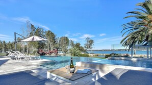 Zona junto a la piscina con una mesa, una botella y vasos, con vistas a un cuerpo de agua y un cielo despejado.