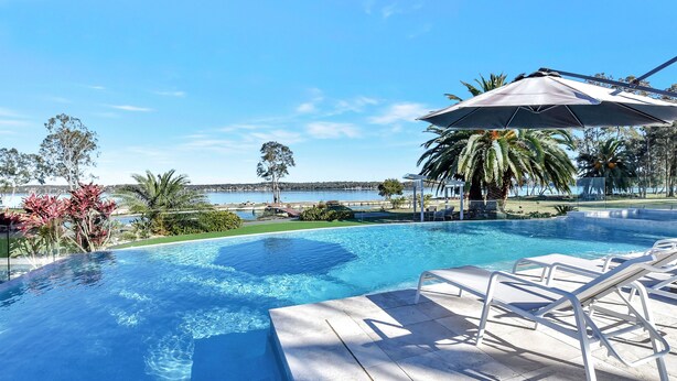 Piscina azul cristalina con vista al mar, rodeada de palmeras y una terraza con sillas reclinables.