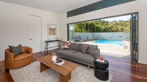 A modern living room with a grey sofa, a brown leather armchair, and a wooden coffee table. There is a glass door leading to a pool area with a view of greenery.