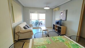 A living room with a sofa, a dining table with chairs, a TV, and a balcony with chairs. - ACV - Torremar-2ª Linea Planta 1 Norte 3H (OROPESA)