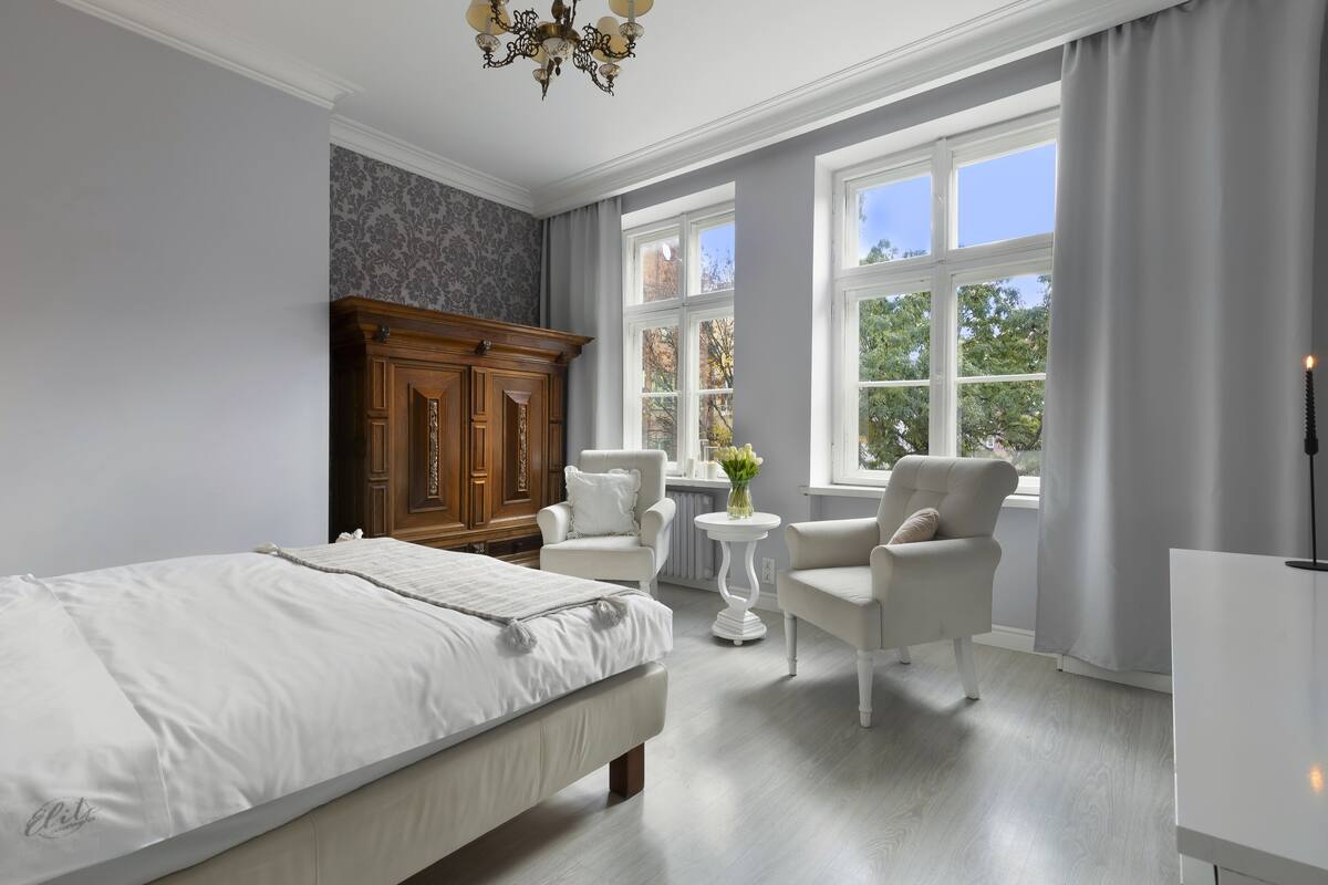A bedroom with a large bed, two white armchairs, a wooden cabinet, and a small table with a vase of flowers.