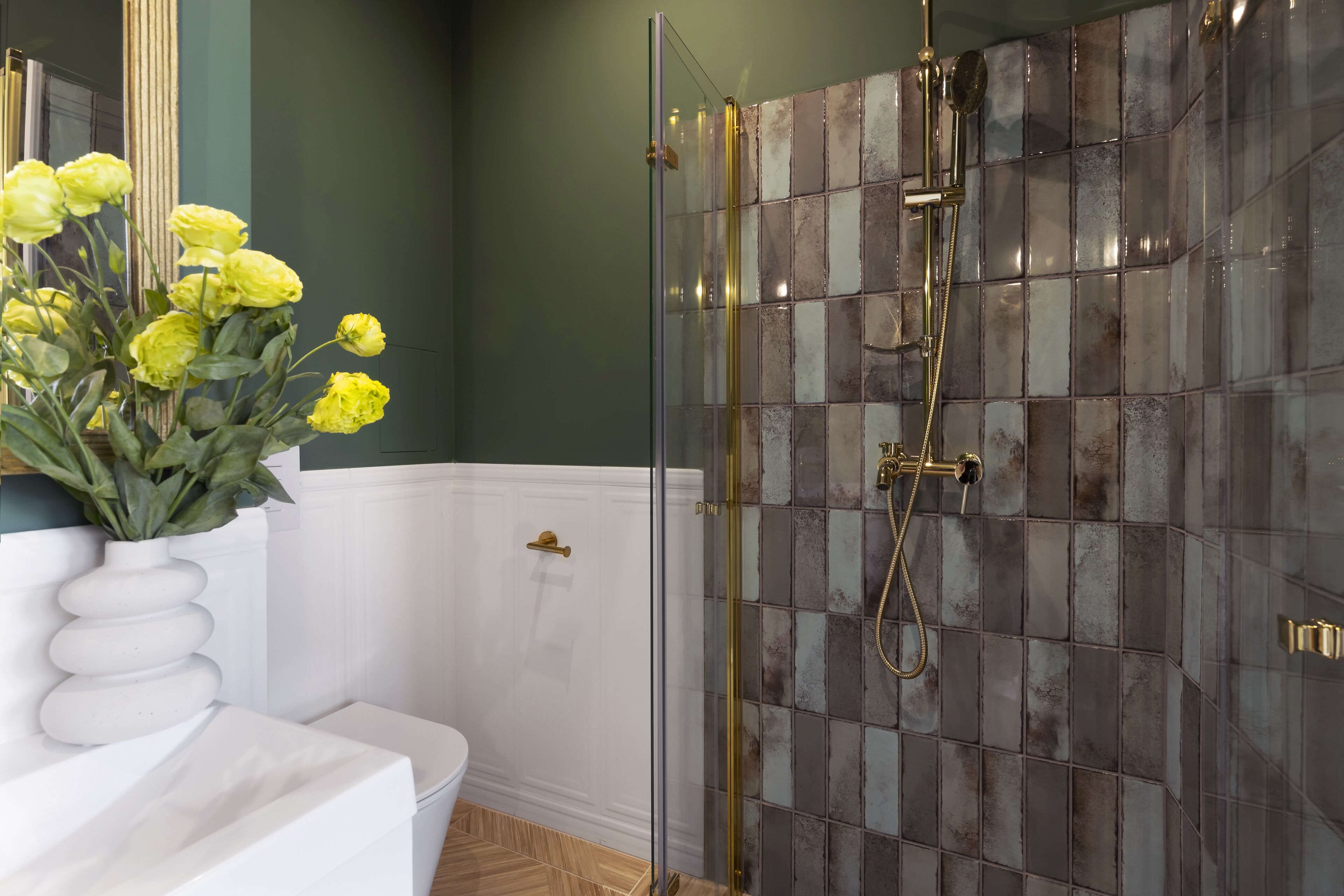 Un baño con ducha de vidrio, inodoro blanco y un florero con flores amarillas sobre un pedestal blanco.