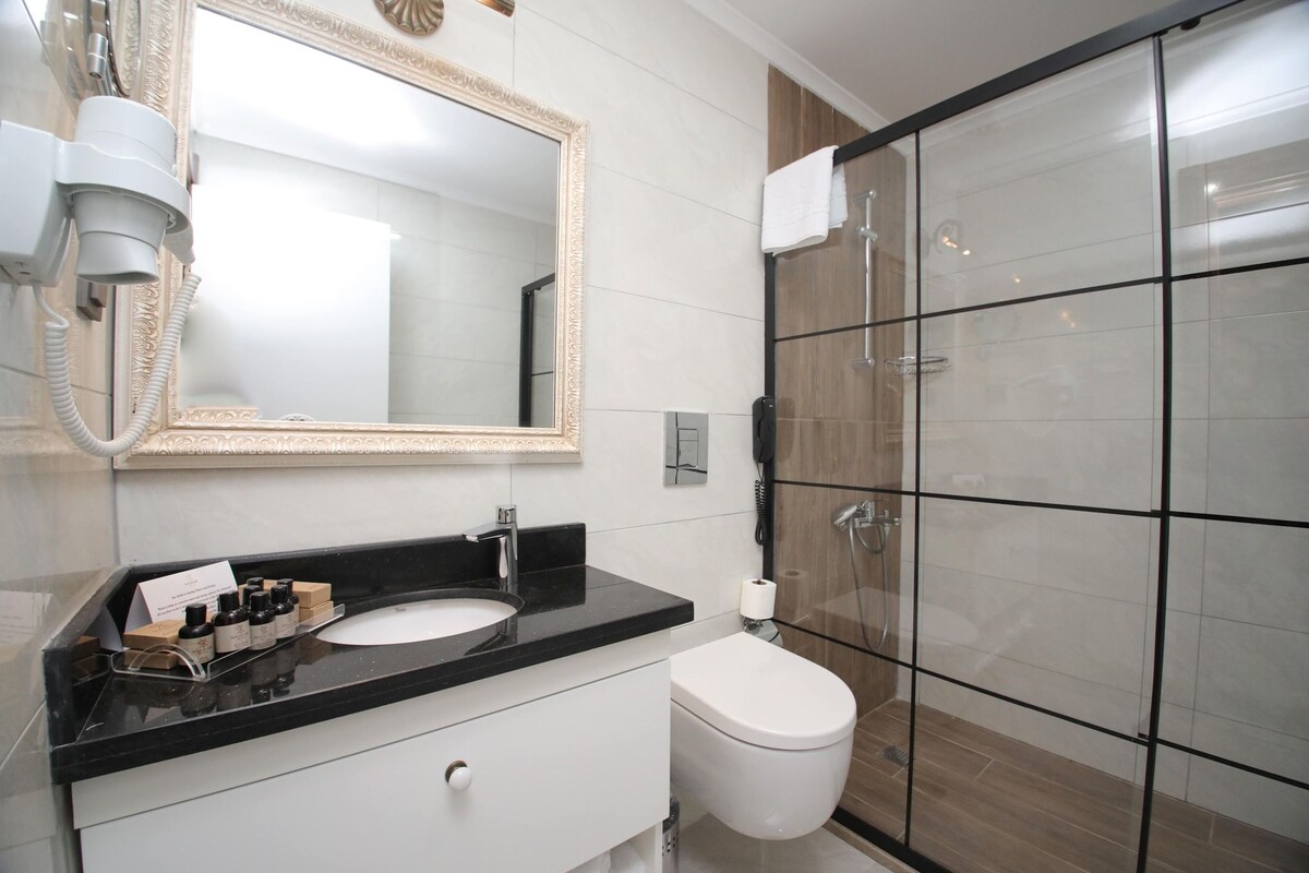 Un baño moderno con encimera negra, lavabo blanco, espejo grande, cabina de ducha con mampara de vidrio y retrete.