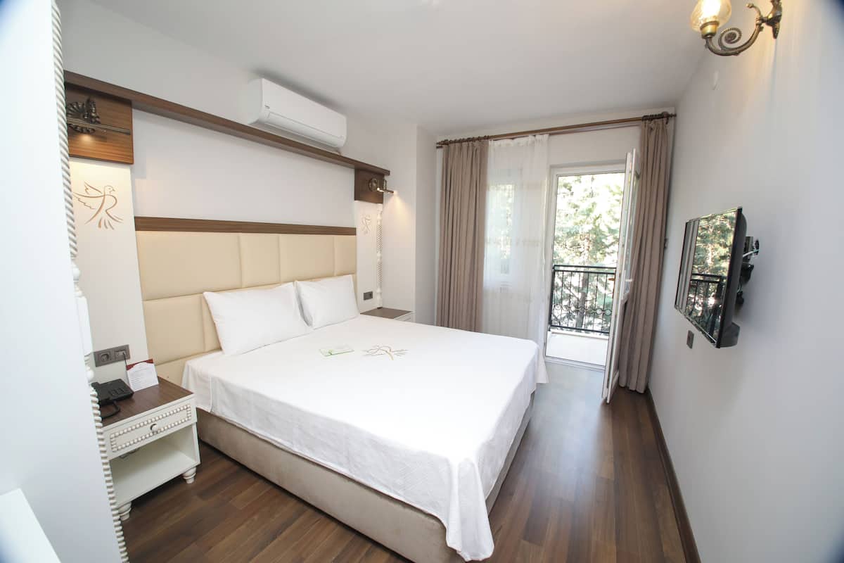 A hotel room with a large bed, a bedside table with a phone and a lamp, a TV mounted on the wall, and a balcony with curtains.