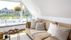 Ein modernes Wohnzimmer mit einem beigefarbenen Sofa, einem kleinen Holztisch und Blick auf eine grüne Landschaft durch große Fenster.