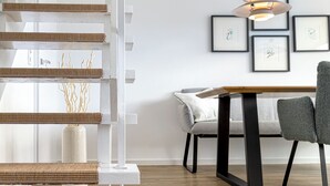 Ein modernes Interieur mit einer Holztreppe, einem Essbereich mit Tisch und Stühlen sowie gerahmten Kunstwerken an der Wand.