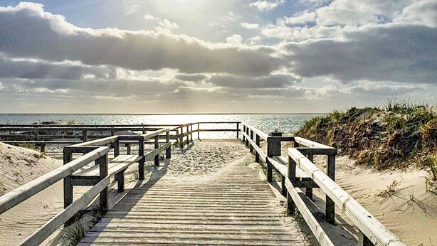Ein hölzerner Steg führt zu einem Strand, der von einem bewölkten Himmel überspanned wird.