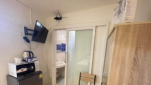 A compact kitchen with a microwave, coffee maker, and a small refrigerator. There is a wooden chair and a sliding glass door leading to a bathroom with a sink. - The Sports Lounge (Pattaya)