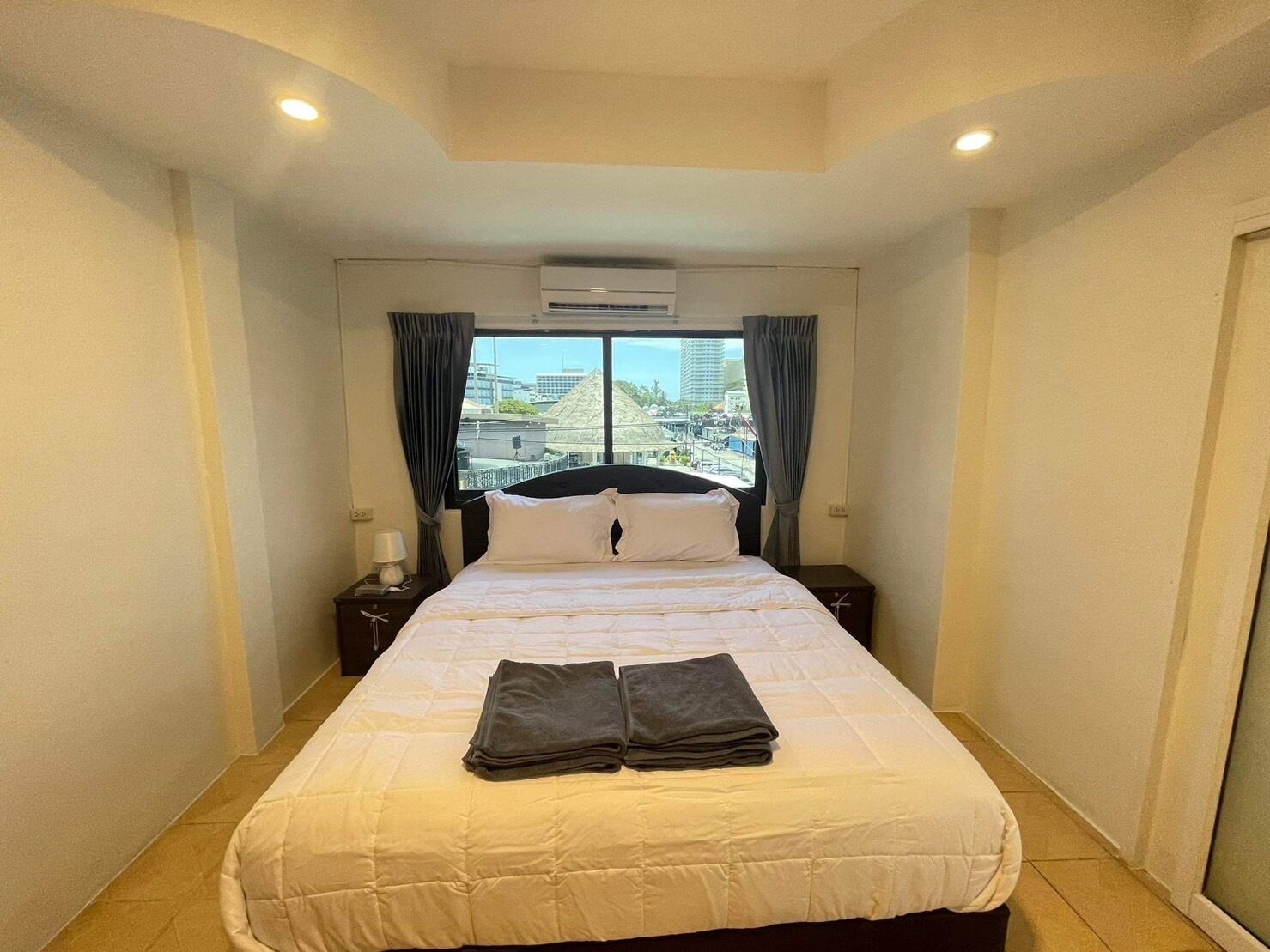 A hotel room with a bed, a nightstand, an air conditioner, and a view of the cityscape through the window.