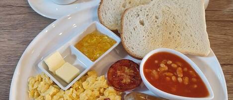A breakfast plate with scrambled eggs, bacon, sausages, toast, beans, tomato, butter, jam, and a cup of coffee.