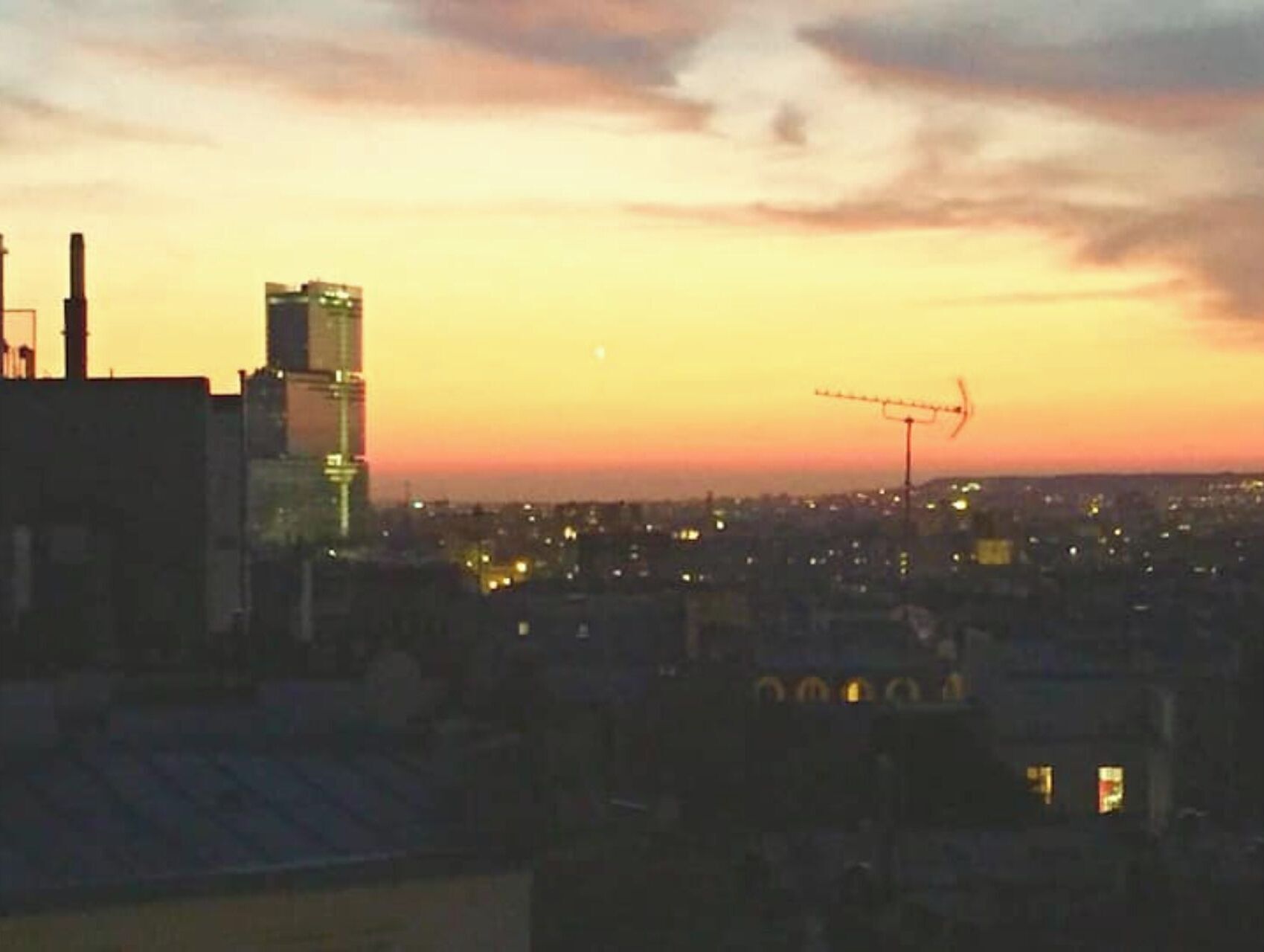 Kaupunkikuva hämärän aikaan, jossa rakennukset ovat valaistuja ja kauempana näkyy rakennustyömaalla oleva nosturi.
