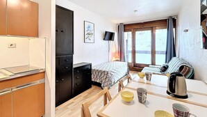 A compact room with a bed, a dining table with chairs, a kitchen area with cabinets, a television, and a window with curtains.