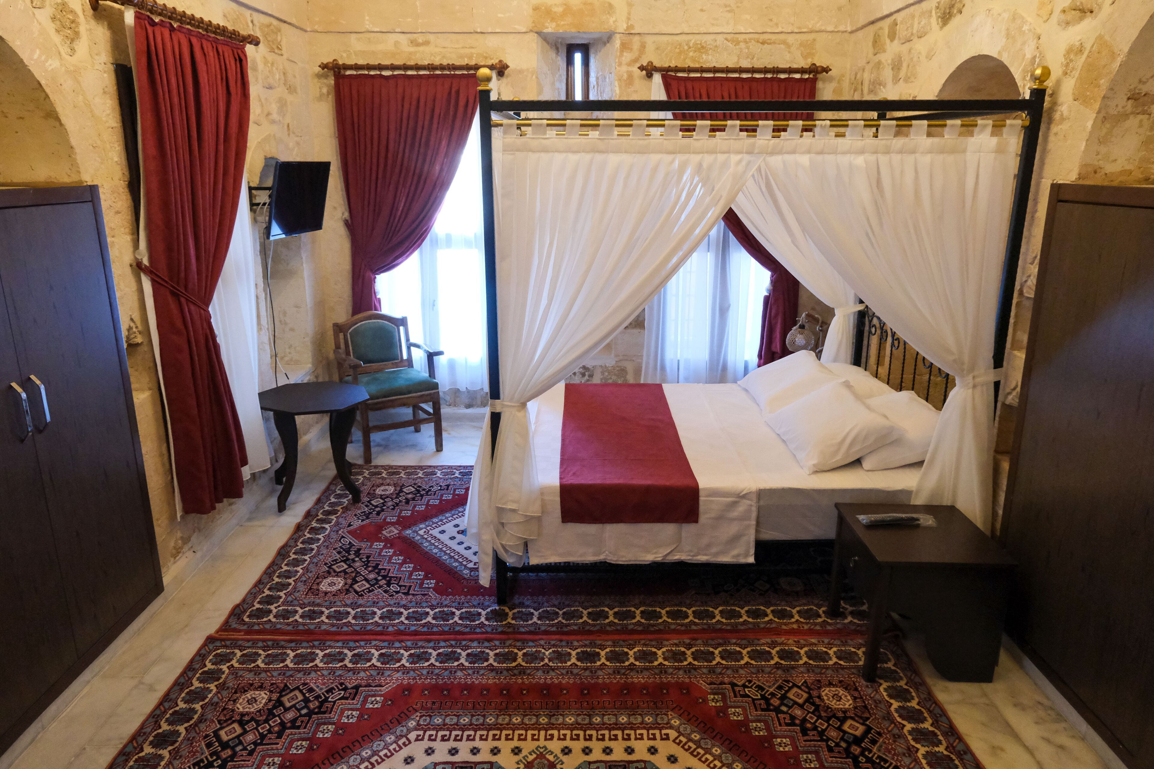A traditional room with a four-poster bed, red and white drapes, a patterned carpet, and stone walls.