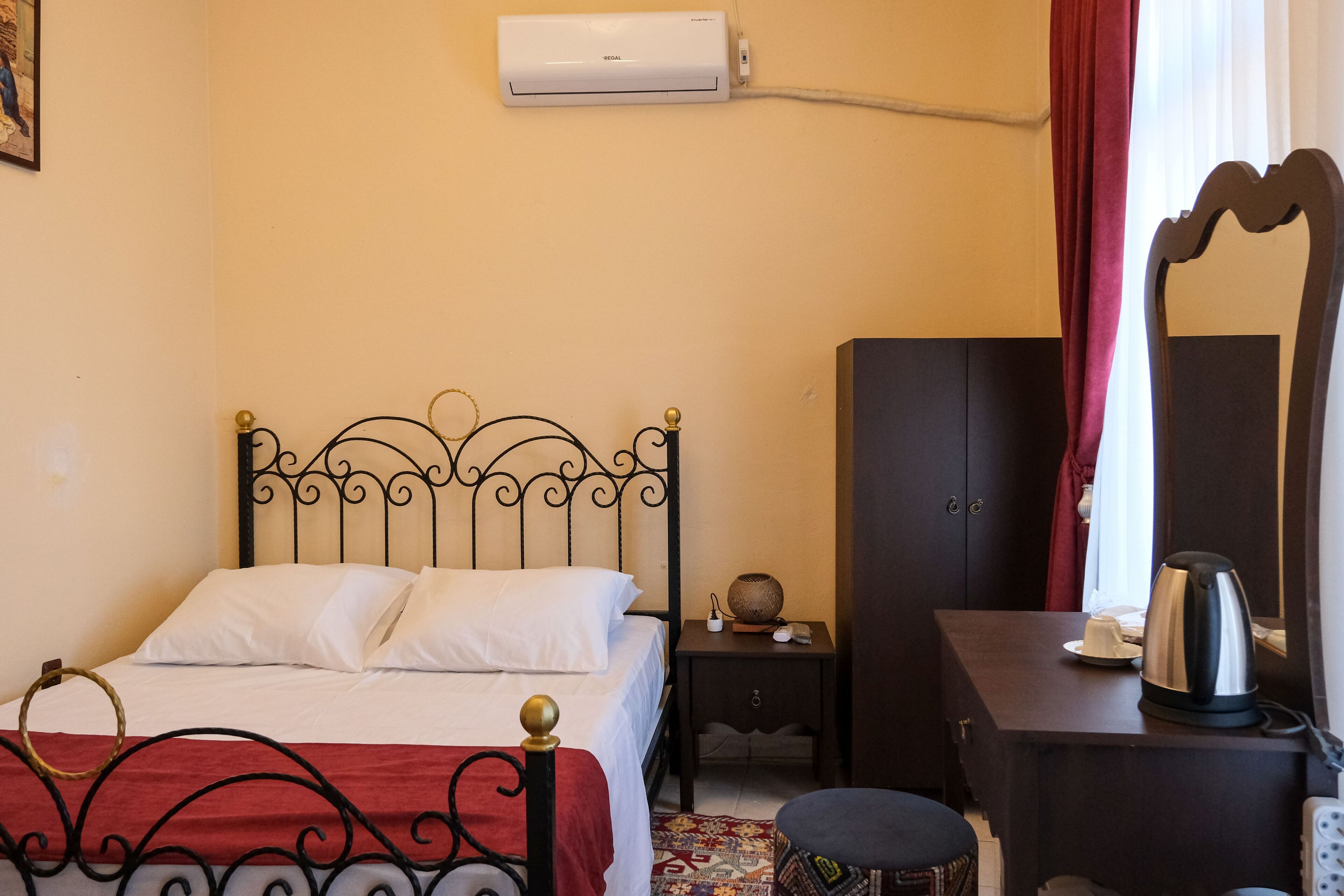 A hotel room with a bed, a small bedside table, a wardrobe, a mirror, and a kettle.