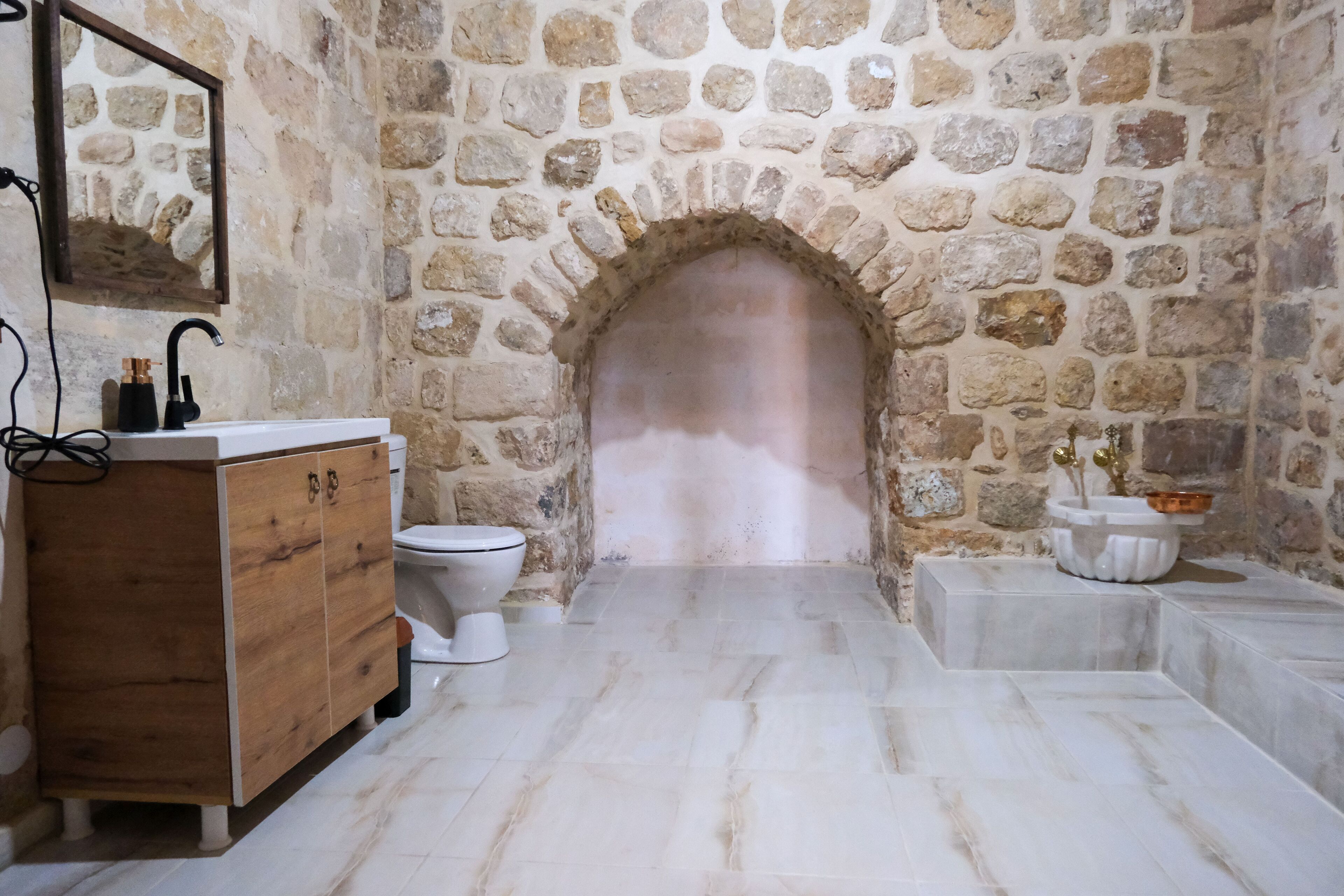 A bathroom with a stone archway, a wooden vanity, a white toilet, and a marble floor.