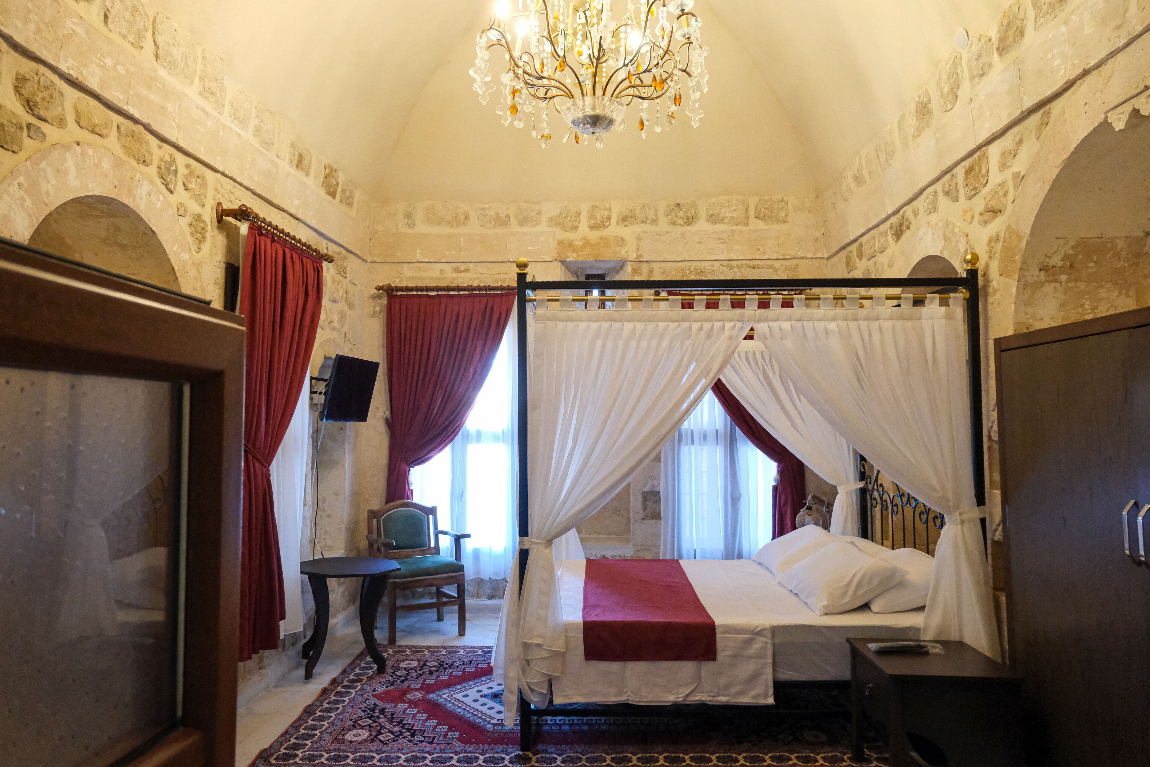 A four-poster bed with white and red bedding, a chandelier, stone walls, and a red and white patterned rug.