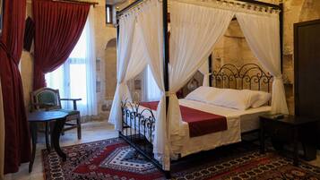 A four-poster bed with white bedspreads and red accents, a wooden chair, a small table, and a patterned rug in a stone-walled room.